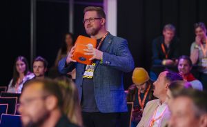 Mann mit Blazer und Namensschild hält orangenes Objekt in einer Veranstaltungshalle mit sitzenden Zuschauern.