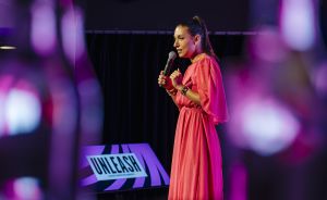 Frau in rosa Kleid spricht mit Mikrofon auf einer Bühne, im Hintergrund ein Schild mit der Aufschrift „UNLEASH".
