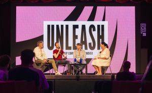 Vier Personen sitzen auf einer Bühne vor einer pinken LED-Wand mit der Aufschrift „UNLEASH" und diskutieren.