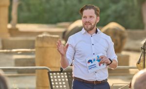 Mann mit Headset-Mikrofon spricht vor Publikum, hält Heft „Sommerempfang" in der Hand, gestikuliert.