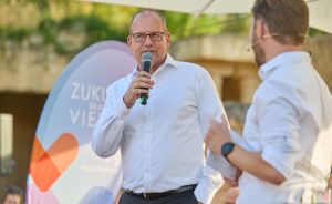 Mann mit Brille spricht in Mikrofon auf Bühne, zweiter Mann im Vordergrund, Banner mit "Zuku… Vie…" im Hintergrund.