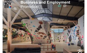 Kletterhalle mit bunten Griffen an Boulderwänden, Holzkonstruktion, Glasdach. Text: „Business and Employment Promotion, Region Hannover"