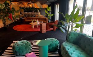 Moderner Loungebereich mit türkisen Samtsofas, gestreiftem Teppich, rotem Bereich mit brauner Couch und tropischen Pflanzen.