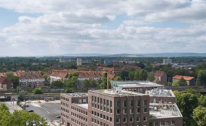 Luftaufnahme eines mehrstöckigen Backsteingebäudes an einer Kreuzung, dahinter Stadtpanorama mit Bäumen und bewölktem Himmel.