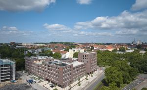 Luftaufnahme einer mittelgroßen Stadt mit mehrstöckigen Backsteingebäuden, breiter Straße, Bäumen und blauem Himmel mit Wolken.