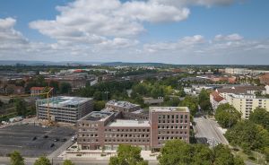 Luftaufnahme einer Stadt mit Bürogebäuden, Baustelle mit Kran, Straßen, Bäumen und Hügeln im Hintergrund bei bewölktem Himmel.