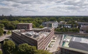 Luftaufnahme eines roten Backsteingebäudekomplexes, umgeben von Bäumen, mit Stadtpanorama im Hintergrund unter bewölktem Himmel.