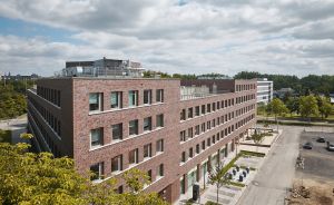 Modernes fünfstöckiges Backsteingebäude mit Flachdach, Parkplatz daneben, Bäume, bewölkter Himmel, Stadtumgebung.