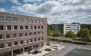 Modernes Backsteinbürogebäude mit Innenhof, dahinter Straße mit Bäumen und weiteres Bürogebäude, bewölkter Himmel.