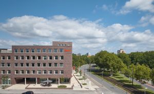 Mehrstöckiges Backsteingebäude mit rotem Logo, daneben eine Straße mit Gleisen, Bäumen und blauem Himmel.