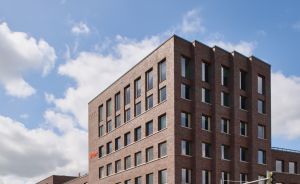 Modernes mehrstöckiges Backsteingebäude an einer Straßenkreuzung unter blauem Himmel mit weißen Wolken.