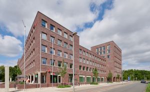 Modernes mehrstöckiges Backsteingebäude an einer Straßenkreuzung, bewölkter Himmel, junge Bäume auf dem Gehweg.