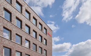 Mehrstöckiges Backsteingebäude mit Außenterrasse, blauem Himmel mit Wolken und weiteren Gebäuden im Hintergrund.
