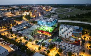 Luftaufnahme einer beleuchteten Stadt bei Dämmerung mit einem markanten, farbenfrohen Gebäude in Blau, Grün und Rot im Zentrum.