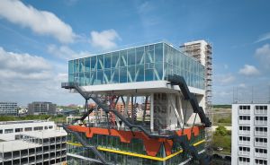 Modernes Glasgebäude mit orangefarbenen Stahlträgern und außenliegenden Treppen, umgeben von Baustellen und Stadtgebäuden.