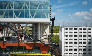 Baustelle mit modernem Glasgebäude und farbigen Elementen neben einem weißen Bürogebäude unter blauem Himmel.
