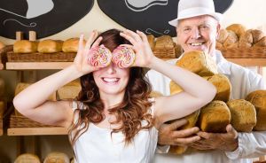Junge Frau hält zwei Cupcakes vor die Augen, älterer Mann in weißer Kochuniform hält Brote in einer Bäckerei.