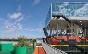 Modernes Gebäude mit Glasfassade, bunten Dachflächen in Orange und Grün, Metalltreppen und blauem Himmel.