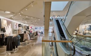 Modebekleidungsgeschäft mit Kleiderständern, Deckenbeleuchtung und einer Rolltreppe im Vordergrund.