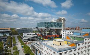 Luftaufnahme einer Stadtlandschaft mit modernem Glasgebäude auf Stahlkonstruktion, umgeben von Wohnblöcken und Bäumen.