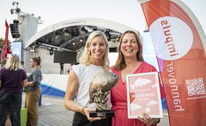 Zwei Frauen stehen vor einer Bühne. Eine hält eine Trophäe, die andere ein Schild mit „Bester Pitch".