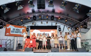 Gruppe von Personen auf einer beleuchteten Bühne mit Banner „Die Höhle der Karpfen", klatschen gemeinsam.
