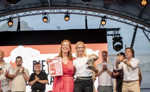 Zwei Frauen auf einer Bühne mit Urkunde und Pokal, umgeben von applaudierenden Personen bei einer Preisverleihung.