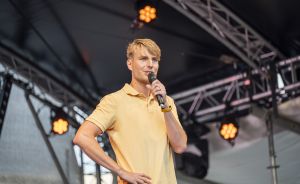 Mann in gelbem Poloshirt hält Mikrofon auf einer Bühne mit Traversen und Scheinwerfern im Hintergrund.