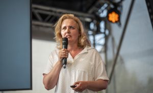 Frau mit blonden Haaren und weißem Hemd hält Mikrofon auf einer Bühne mit Bühnentechnik im Hintergrund.