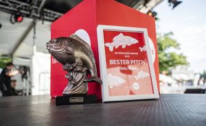 Silberne Fisch-Trophäe neben rotem Schild „Bester Pitch 500,- Euro" auf einem Tisch, im Hintergrund eine Bühne.