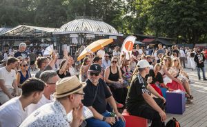 Große Menschenmenge sitzt und steht im Freien bei einem Outdoor-Event, im Hintergrund ein Pavillon und Bäume.