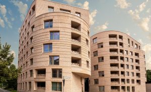 Modernes mehrstöckiges Wohngebäude aus hellem Backstein mit geschwungener Fassade, Balkonen und großen Fenstern, blauer Himmel.