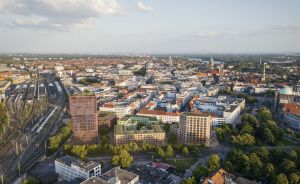 Luftaufnahme einer deutschen Großstadt mit Bahngleisen, Wohn- und Geschäftsgebäuden sowie Kirchturm bei sonnigem Wetter.