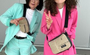 Zwei Frauen posieren lächelnd: eine im türkisen, die andere im pinken Blazer-Shorts-Outfit, beide mit beigen Umhängetaschen.