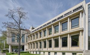 Zweigeschossiges Steingebäude mit Säulenfassade und großen Fenstern, daneben moderner Glasanbau, Baum, Rasenfläche.