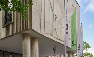 Modernes Steingebäude auf Säulen, grüne Fahnen mit "Hochschule"-Aufschrift, Bäume, Personen am Eingang, blauer Himmel.
