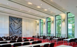 Konferenzsaal mit Stuhlreihen, rotem Teppich, großen Fensterfronten mit Baumblick und abstrakter Wandkunst.