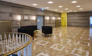 Helles Foyer mit Terrazzoboden, schwarzen Sesseln, Stehlampen, Marmor­wänden und vergoldetem Treppengeländer.