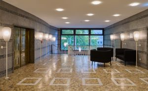 Helle Lobby mit Terrazzoboden, geometrischem Muster, schwarzen Sesseln, weißen Stehlampen und Fensterfront mit Baumblick.