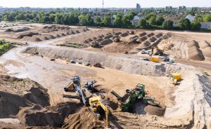 Luftaufnahme einer großen Baustelle mit Erdaushub, Baumaschinen und Baggern, im Hintergrund eine Stadt mit Bäumen.