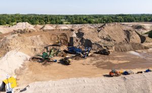 Luftaufnahme einer Sandgrube mit Baumaschinen, Förderbändern, Erdhügeln und Bäumen im Hintergrund.