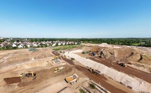 Luftaufnahme einer Baustelle mit mehreren gelben Baumaschinen auf sandigem Untergrund, im Hintergrund Wohnhäuser und Bäume.