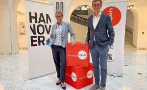 Zwei Personen stehen vor Hannover- und hannoverimpuls-Bannern neben roten Würfeln mit hannoverimpuls-Logo.