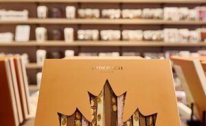Goldene Geschenkbox mit Ahornblatt-Ausschnitt, dahinter sichtbare Schokoladenriegel, im Hintergrund Ladenregale.