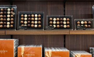 Holzregal mit Läderach-Schokoladenboxen: oben Pralinenschachteln, unten orange und weiße Geschenkboxen mit Schleife.