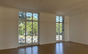 Leerer Raum mit Holzboden, weißen Wänden, zwei großen Sprossenfenstern mit Blick auf Bäume und Straße.
