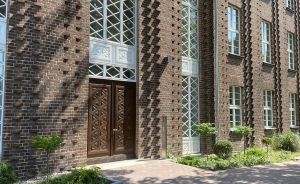 Braunes Backsteingebäude mit großen Sprossenfenstern, verzierten Holzdoppeltüren und gepflastertem Eingangsweg mit Begrünung.