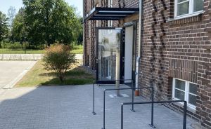 Fahrradständer aus Metall vor einem Backsteingebäude mit Glaseingang und Vordach, gepflasterter Vorplatz, Bäume im Hintergrund.