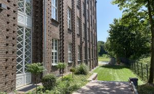 Backsteingebäude mit verzierten Fassadenstreifen, gepflasterter Weg, Rasenfläche und Bäume bei Sonnenschein.