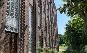 Backsteingebäude mit Zickzack-Ornamentik, gepflasterter Weg, Bäume und blauer Himmel an einem sonnigen Tag.